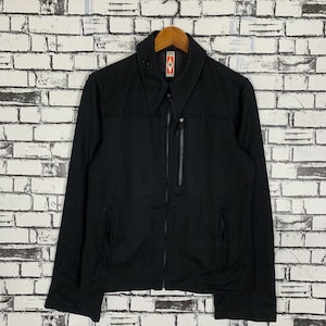 Puede incluir: Una chaqueta negra con cremallera, cuello y dos bolsillos en la parte delantera. La chaqueta tiene una pequeña etiqueta en el pecho que dice "Evisu".