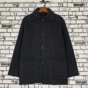 Puede incluir: Un abrigo de lana gris carbón con cuello clásico, tres botones y dos bolsillos delanteros. El abrigo está colgado en una percha de madera contra una pared de ladrillos blancos.