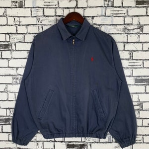 Puede incluir: Una chaqueta azul marino con cremallera con un logotipo de jugador de polo bordado en rojo en el pecho izquierdo. La chaqueta tiene una cintura y puños de canalé.