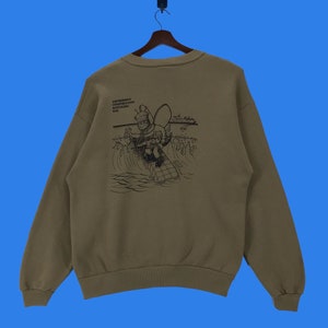 May include: A brown sweatshirt with a black graphic print of a cartoon character fishing on a surfboard. The text "AMPHIBIOUS CONSTRUCTION INTERNATIONAL DIVE" is printed above the graphic.
