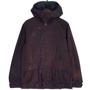 Puede incluir: Una chaqueta con capucha de color morado oscuro con costuras rojas y forro a cuadros. La chaqueta tiene dos bolsillos grandes en la parte delantera y cierre de botones.