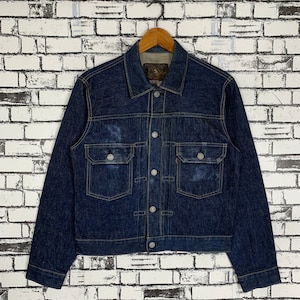 Puede incluir: Chaqueta vaquera azul oscuro con dos bolsillos en el pecho y dos bolsillos laterales. La chaqueta tiene botones plateados y un cuello clásico. La chaqueta está colgada de una percha de madera contra una pared de ladrillos blancos.