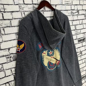 Puede incluir: Una sudadera con capucha gris oscuro de aspecto vintage. Presenta un gran parche bordado en la espalda con un avi&oacute;n y el texto "Air Force 9". Hay un parche m&aacute;s peque&ntilde;o en la manga izquierda con el texto "Air Force" y una estrella.