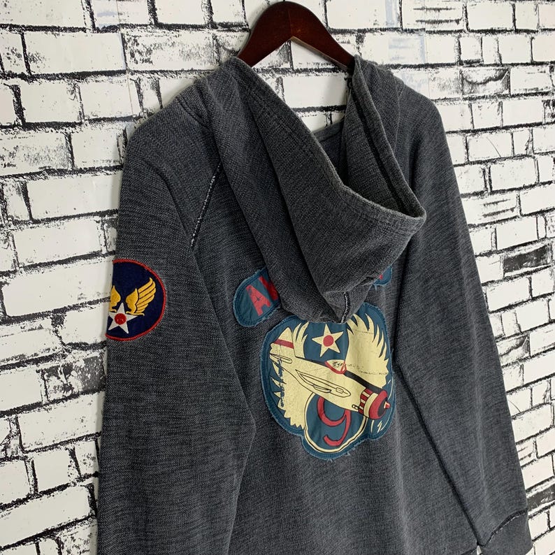Puede incluir: Una sudadera con capucha gris oscuro de aspecto vintage. Presenta un gran parche bordado en la espalda con un avi&oacute;n y el texto "Air Force 9". Hay un parche m&aacute;s peque&ntilde;o en la manga izquierda con el texto "Air Force" y una estrella.