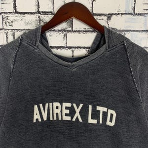Puede incluir: Una sudadera con capucha negra con el texto "AVIREX LTD" en letras blancas.