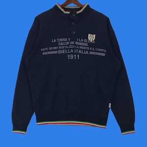 Puede incluir: Una sudadera azul marino con cuello y puños a rayas blancas, rojas y verdes. La sudadera tiene un logotipo blanco con el texto "LA CORSA V LA GLUA FACCIO UN BRINDISI. FARE SPORT RIVITALIZZA LA MENTE E IL CORPO. BIELLA ITALIA 1911" y un pequeño logotipo de Fila.