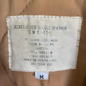 May include: A white tag with black text detailing the features of a jacket. The tag reads "Jacket Flyer's Cold Weather CWU-55", "AV-100PAY 8338 NYCPR", "C100-81-50EW", "AVIREXTO", "Shell 100% Nylon", "Lining 100% Nylon", "Insulation 100% Polyester", "Dry Clean Only". A white tag with the letter "M" is visible below the larger tag.