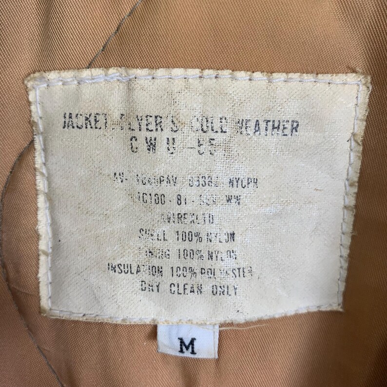 May include: A white tag with black text detailing the features of a jacket. The tag reads "Jacket Flyer's Cold Weather CWU-55", "AV-100PAY 8338 NYCPR", "C100-81-50EW", "AVIREXTO", "Shell 100% Nylon", "Lining 100% Nylon", "Insulation 100% Polyester", "Dry Clean Only". A white tag with the letter "M" is visible below the larger tag.