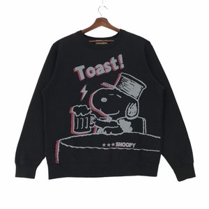 Peut inclure: Sweat-shirt noir avec un imprimé graphique de Snoopy portant un chapeau haut de forme et tenant une chope de bière. Le texte "Toast!" est imprimé au-dessus de Snoopy et le texte "***Snoopy" est imprimé en dessous.