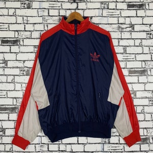 Puede incluir: Una chaqueta vintage Adidas de color azul, rojo y blanco con cierre de cremallera y el logotipo de Adidas en el pecho.