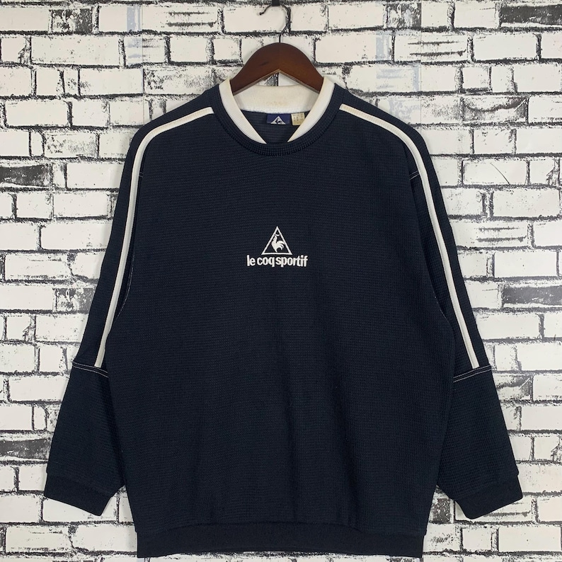 Puede incluir: Una sudadera azul marino con rayas blancas en las mangas y un logotipo bordado en blanco que dice "le coq sportif" con un gallo dentro de un tri&aacute;ngulo.