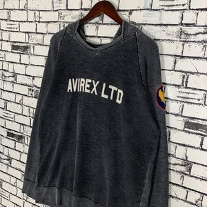 Puede incluir: Una sudadera con capucha negra con un aspecto descolorido. La sudadera tiene el texto "AVIREX LTD" en letras blancas en la parte delantera y un peque&ntilde;o parche amarillo y negro en la manga izquierda.