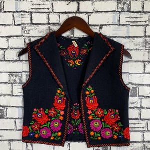 Puede incluir: Un chaleco negro con vibrante bordado floral en rojo, rosa, verde y amarillo. El chaleco presenta un cuello con muescas y está ribeteado con un borde cosido en rojo. El diseño floral se concentra en la parte delantera y superior de la espalda.