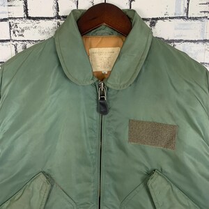 May include: A green bomber jacket with a zipper closure and a patch on the right sleeve. The jacket has a collar and a quilted lining.