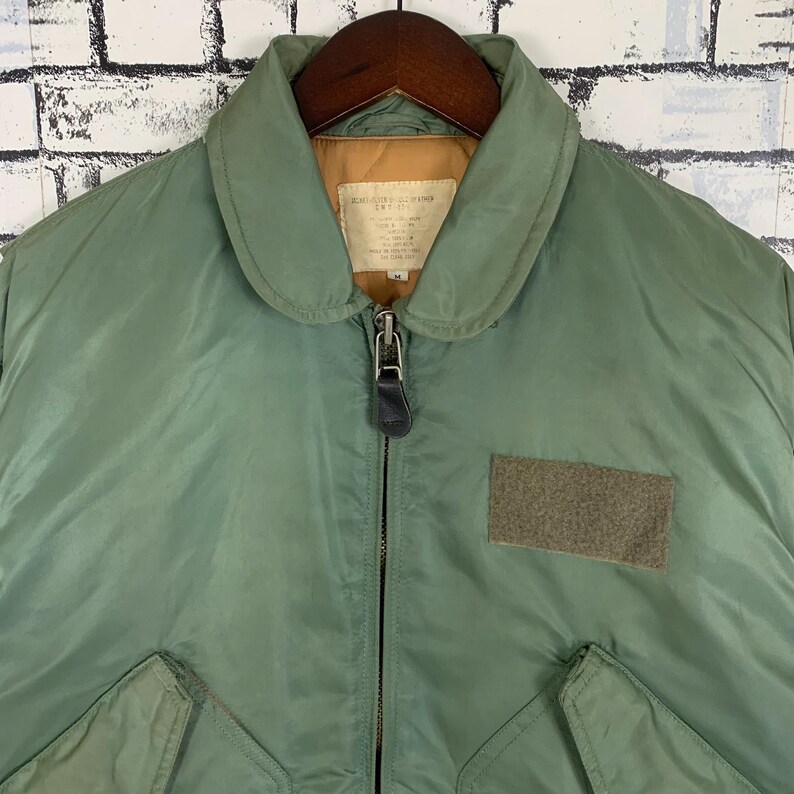 May include: A green bomber jacket with a zipper closure and a patch on the right sleeve. The jacket has a collar and a quilted lining.