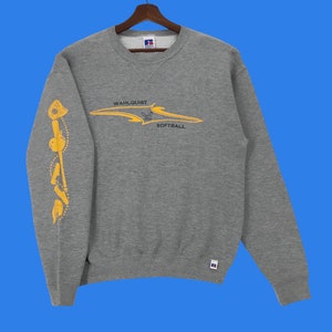 Puede incluir: Sudadera gris con un gráfico amarillo en la parte delantera que dice "Wahlquist Softball". La manga izquierda tiene un gráfico amarillo de un jugador de softbol con un bate y una pelota.