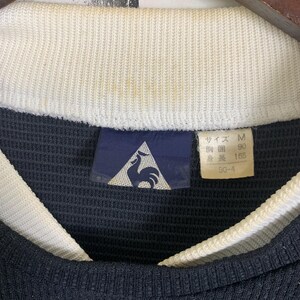 Puede incluir: Una etiqueta de talla negra y blanca con un logotipo de gallo. La etiqueta dice "Talla M, 90, 165, 50-4".