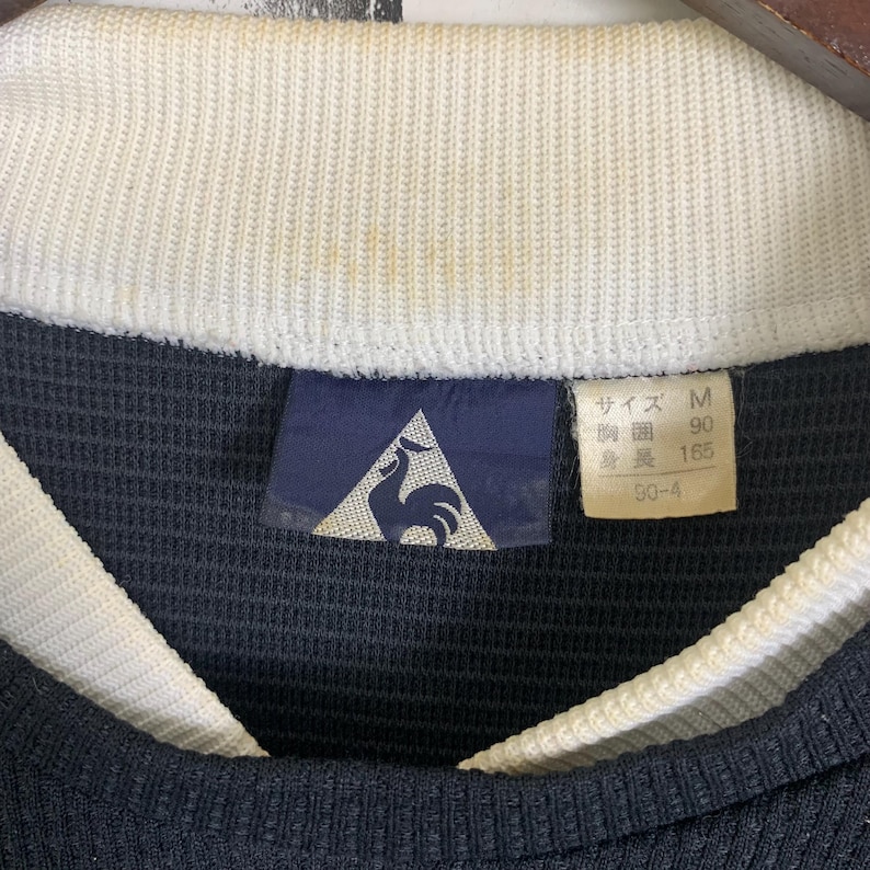 Puede incluir: Una etiqueta de talla negra y blanca con un logotipo de gallo. La etiqueta dice "Talla M, 90, 165, 50-4".