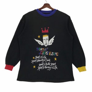 May include: Black long-sleeve sweatshirt with a colorful graphic design. The design includes a face with wings, a crown, stars, and text that reads "SPORTS JCECAS ELBA AC". The cuffs are red and yellow, and the collar is blue.