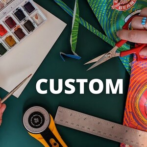 May include: A green cutting mat with a ruler, rotary cutter, scissors, paintbrushes, watercolor paints, a sponge, and a bag of coffee beans. The text "CUSTOM" is written in white letters on the mat.