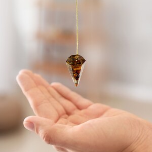 May include: A brown and gold crystal pendulum hanging from a gold chain over a person's open hand.