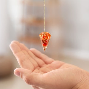 May include: A gold chain with a red and orange crystal pendulum hanging from it. The pendulum is suspended over an open hand.