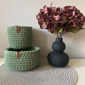 Könnte beinhalten: Zwei grüne gehäkelte Körbe, die übereinander auf einem runden grauen Platzdeckchen gestapelt sind. Im Hintergrund steht eine schwarze Vase mit einem Trockenblumenarrangement.