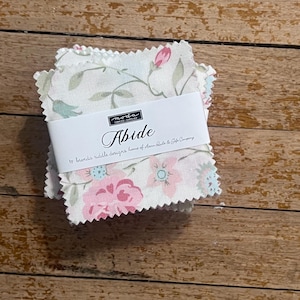 May include: A stack of fabric squares with a floral pattern in pink, blue, and green on a white background. The fabric is wrapped with a white label that reads "Abide" and "moda fabrics supply". The squares are on a wooden surface.