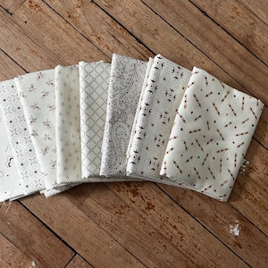 May include: A collection of folded fabric squares in various patterns, including floral, paisley, and geometric designs. The fabrics are primarily white with accents of brown and gray, arranged on a wooden surface.