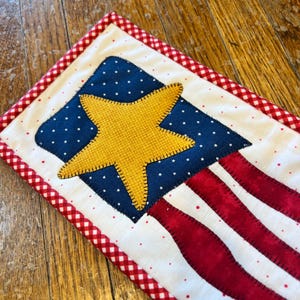 May include: A small quilted American flag with a large yellow star in the center. The flag is surrounded by a red and white gingham border.