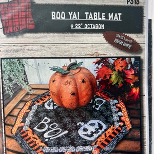 May include: A black and white table mat with a spiderweb design and the word "BOO!" in white. The mat is surrounded by a black and orange border with white polka dots. A pumpkin with a spiderweb design is in the center of the mat. The mat is 22 inches in diameter and is an octagon shape.