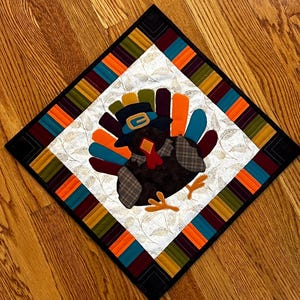 May include: A quilted Thanksgiving-themed table topper featuring a turkey wearing a pilgrim hat. The turkey is brown with colorful tail feathers. The square topper has a white center with a gold leaf pattern, framed by a striped border in fall colors and a black border.