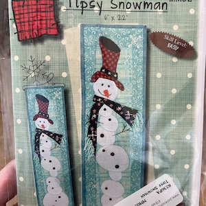 May include: A quilting pattern for a whimsical snowman with a red and white hat, a scarf, and a blue background. The pattern is titled "Tipsy Snowman" and is 6 inches by 22 inches. The skill level is listed as "Easy".