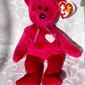 May include: A pink beanie baby bear with a white heart on its chest and a pink ribbon around its neck. The bear has black eyes and a black nose. The bear is sitting on a white background. The bear has a Ty tag attached to its ear.