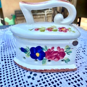 May include: A white ceramic iron shaped box with a floral design. The iron has a handle and a lid. The flowers are painted in shades of pink, blue, and green.