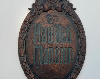 May include: A dark brown, oval-shaped sign with a green, ornate frame. The sign reads "The Haunted Mansion" in a stylized font.