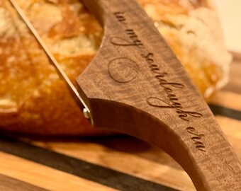 May include: A wooden bread lame with a stainless steel blade, resting on a wooden cutting board. The lame is engraved with the words "in my sourdough era". A loaf of bread is in the background.