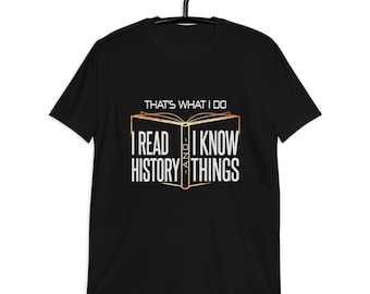 Puede incluir: Camiseta negra con un gráfico dorado y blanco de un libro abierto. El texto en la camiseta dice "That's what I do. I read history and I know things."