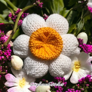 May include: A white crocheted flower with a yellow center. The flower is surrounded by other flowers and greenery.