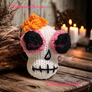 May include: A white knitted skull with black, pink, and orange accents. The skull has black eyes, a stitched mouth, and orange hair. The image includes the text "Stephanie Stitches".