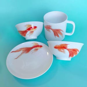 Puede incluir: Un juego de vajilla de porcelana blanca con diseños de peces dorados pintados a mano. El juego incluye una taza, un cuenco y un platillo.