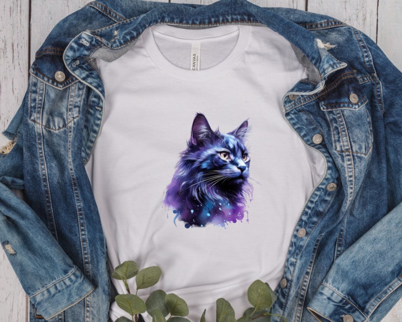 May include: White t-shirt featuring a watercolor illustration of a blue and purple cat. The cat's fur has a galaxy-like design. The shirt is displayed on a denim jacket.