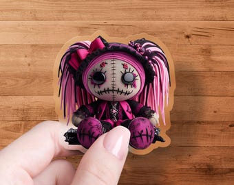 May include: A sticker of a gothic doll with a stitched face, large black eyes, and pink hair. The doll wears a black hat with a pink bow and a pink and black outfit. The doll's legs are a deep pink color.