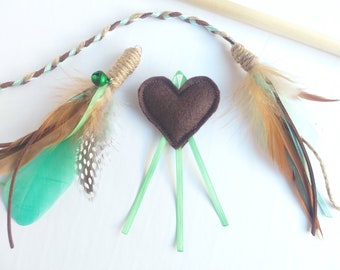 May include: A cat toy with three different attachments: a brown felt heart with green ribbon streamers, a brown and tan feather with a green bell, and a brown and tan feather with a green ribbon.