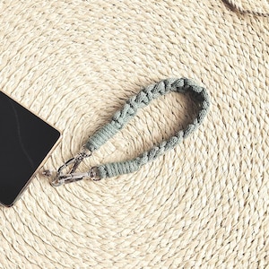 May include: A black smartphone with a silver camera module and a light green macrame wrist strap. The phone rests on a round, woven placemat with a beige rope. The background is a white marble surface.