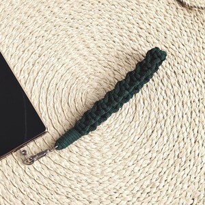 Puede incluir: Un smartphone negro con un marco dorado y una correa de muñeca de macramé verde oscuro. El teléfono y la correa están sobre un salvamanteles redondo y tejido. También hay una fina cuerda beige.
