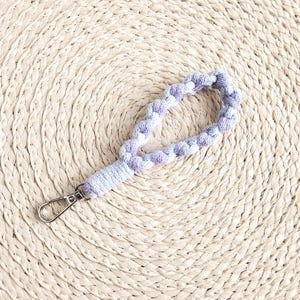 May include: A braided wrist strap in shades of lavender and white, attached to a silver-colored clasp. The strap is resting on a round, woven placemat made of natural fibers. A length of rope is also visible.