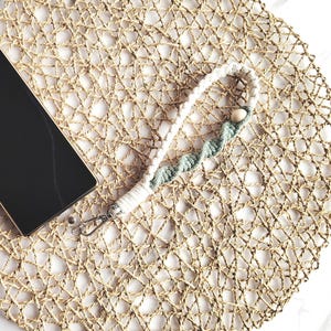 May include: A black smartphone with a silver camera module rests on a round, woven placemat. A white and teal macrame wrist strap with wooden beads is attached to the phone. The placemat is made of natural fibers.