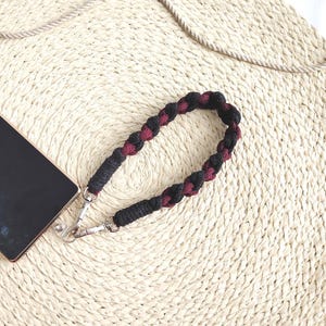 May include: A black smartphone with a gold-colored camera module and a black and burgundy wrist strap. The strap is made of braided cord and has silver-colored metal clasps. The phone rests on a round, woven placemat.