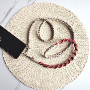 May include: A macrame phone strap in beige and red-brown. The strap is attached to a black phone case with silver hardware. The phone and strap are on a woven beige placemat, set against a white marble surface. The strap is a stylish accessory.